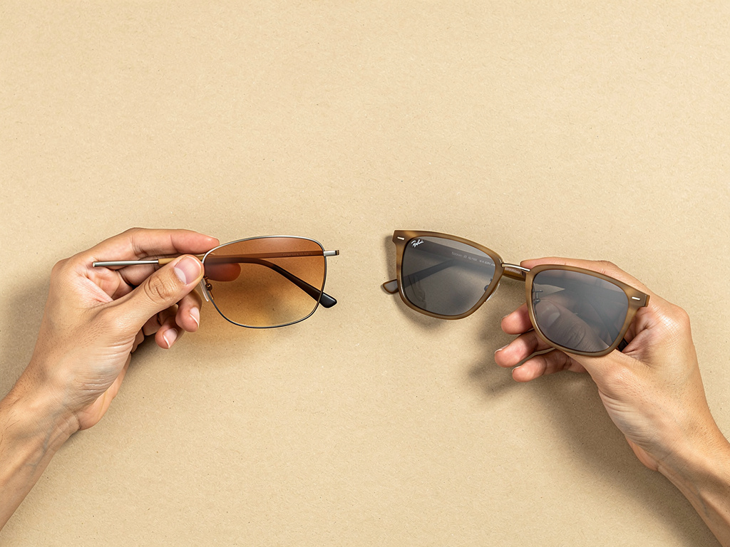 Hands swapping one pair of sunglasses for another on a neutral sand-toned surface.